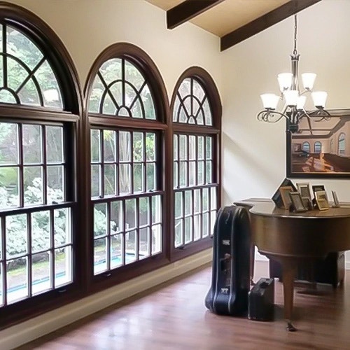 A cozy interior featuring arched brown windows, a grand piano, and a chandelier, surrounded by greenery.