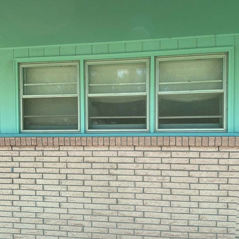 Three teal-framed jalousie (louvered) windows above a beige brick exterior wall on a mid-century home