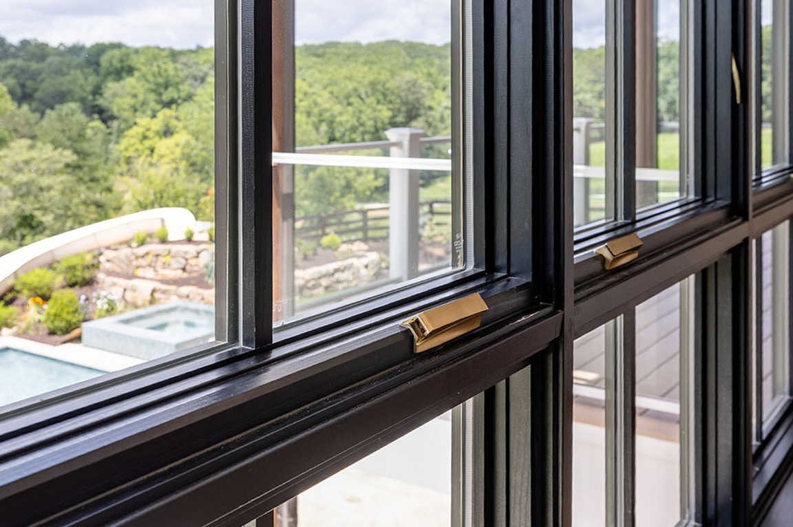 Close-up view of modern black frame windows with brass handles, overlooking a landscaped backyard with a pool and lush greenery.
