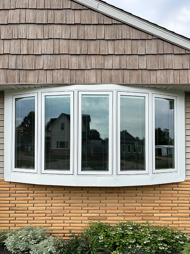 Close-up of a white bow window with five panels installed on a home exterior featuring tan siding and brick accent.