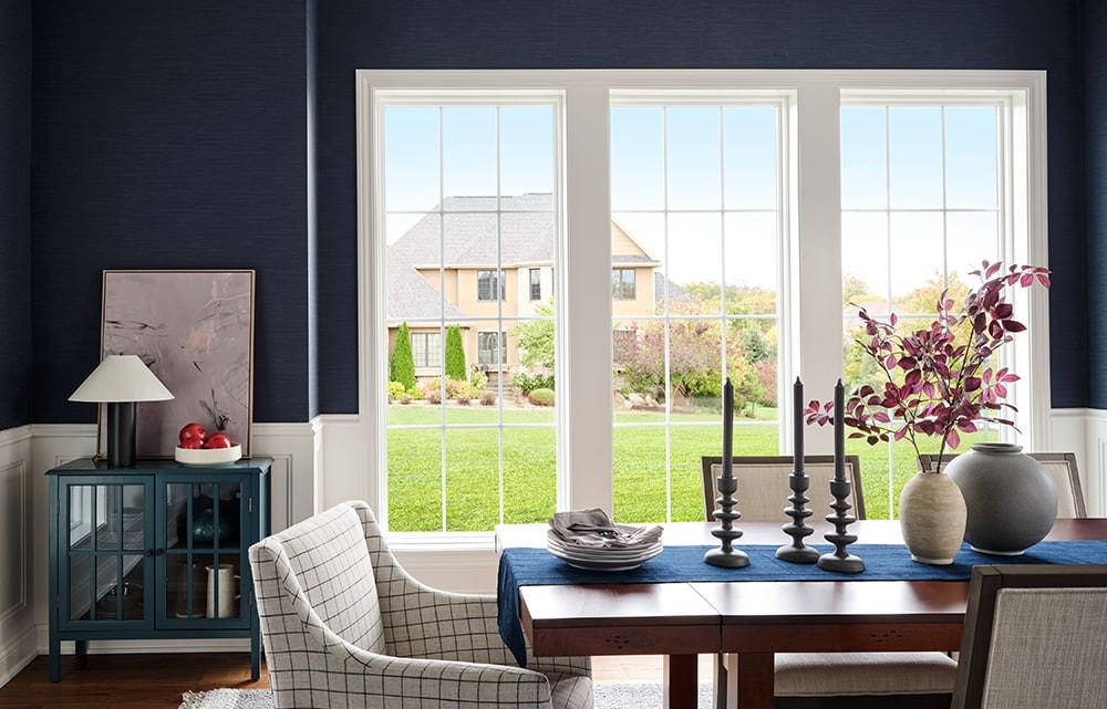 White vinyl windows on a dark blue wall looks out to a large green backyard and houses in the distance.