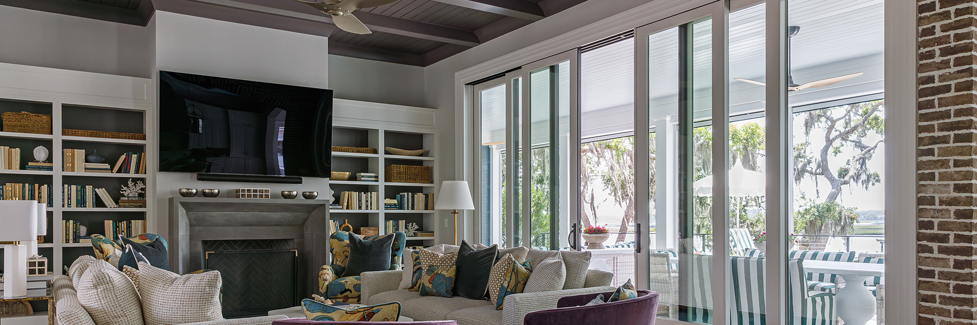 Modern living room featuring large sliding glass doors, cozy seating area with patterned cushions, built-in bookshelves, and a wall-mounted TV above a fireplace.