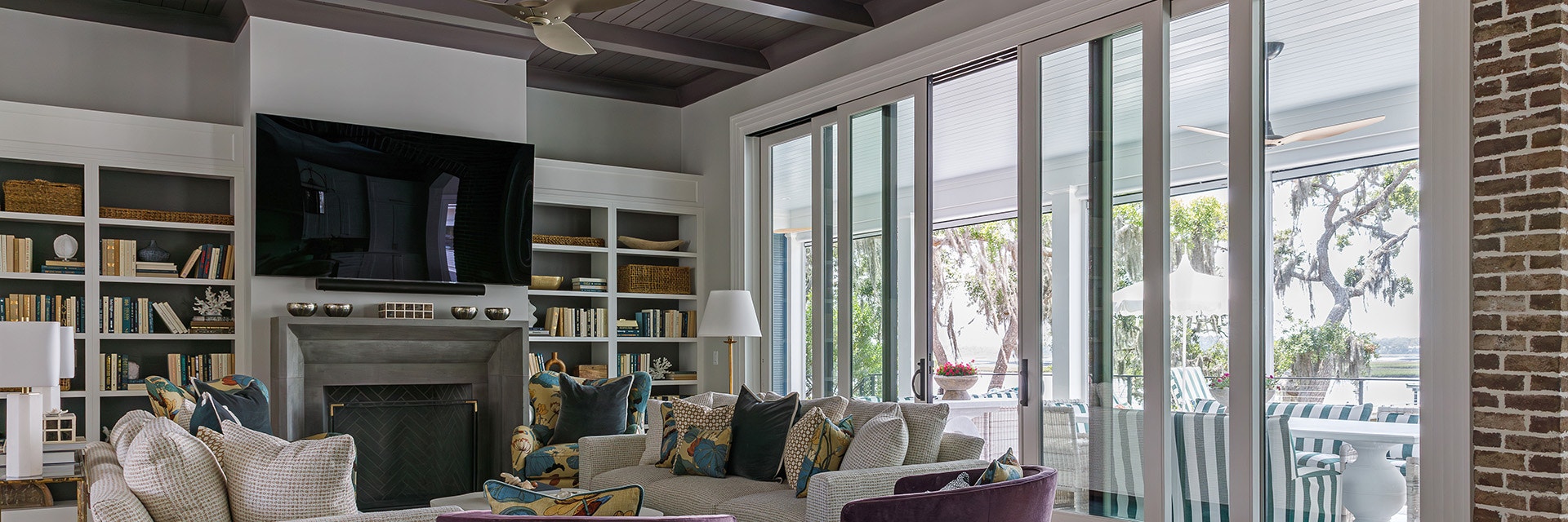 Modern living room featuring large sliding glass doors, cozy seating area with patterned cushions, built-in bookshelves, and a wall-mounted TV above a fireplace.