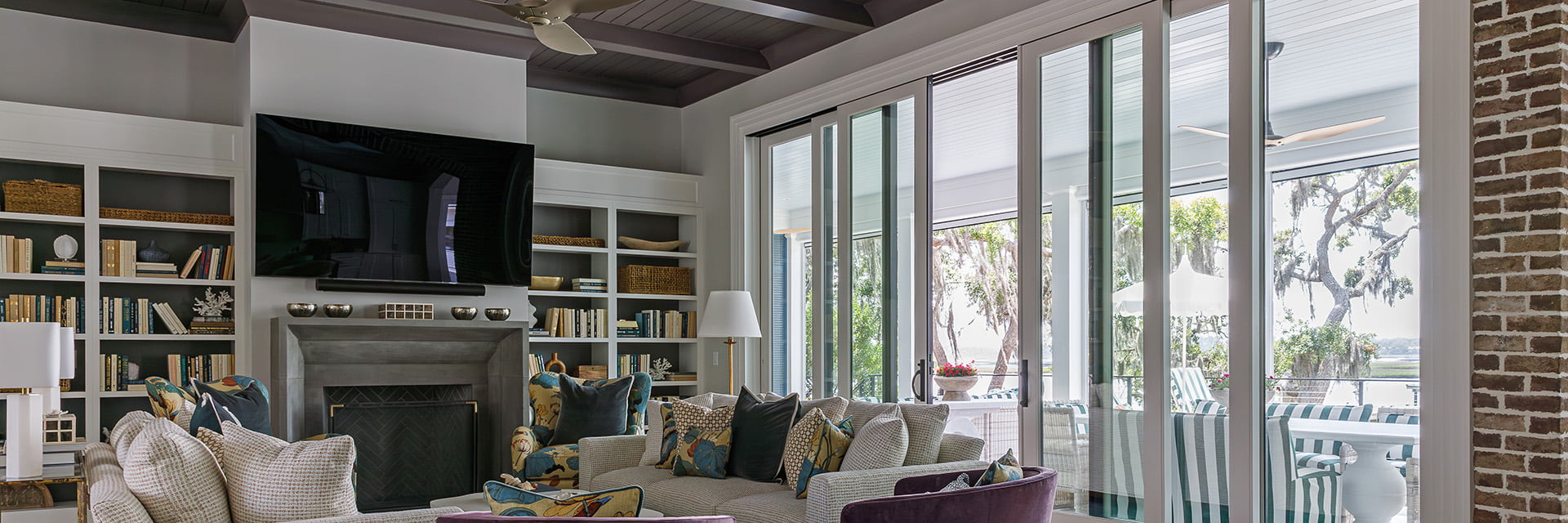 Modern living room featuring large sliding glass doors, cozy seating area with patterned cushions, built-in bookshelves, and a wall-mounted TV above a fireplace.