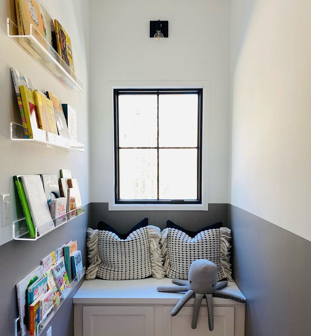 A small nook in a bedroom features shelves of books on the wall and a window seat below a black casement window.