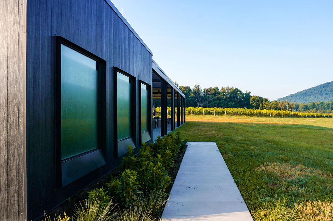 Three large extruded fixed windows on a black wood building with a sidewalk along the exterior and lots of green grass.