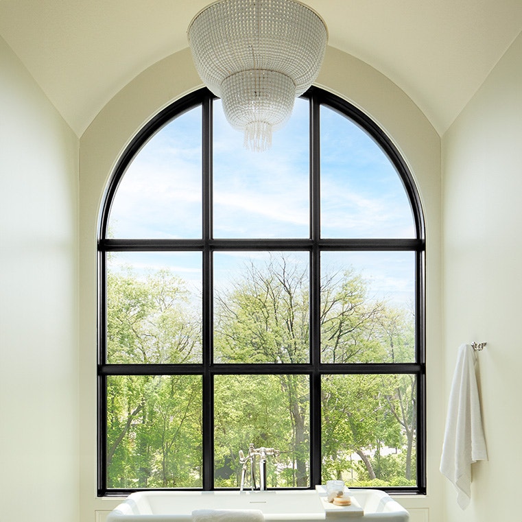 A bright bathroom featuring a large arched window with traditional grilles, overlooking greenery, and a freestanding bathtub.