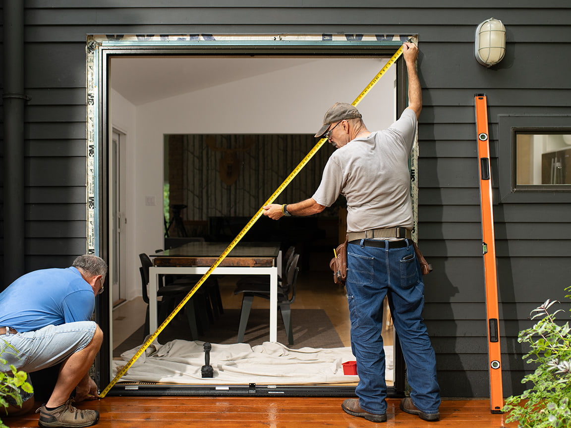 Two men are measuring the frame opening hole in a new home so they can install a new sliding patio door.