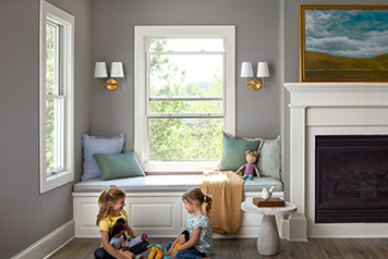 two little girls are sitting on the floor in front of a window seat, that sits below a white double-hung window.