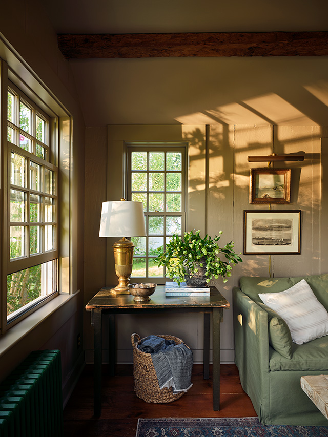 A cozy Cape Cod living room with double-hung windows, a green sofa and a side table with a lamp.