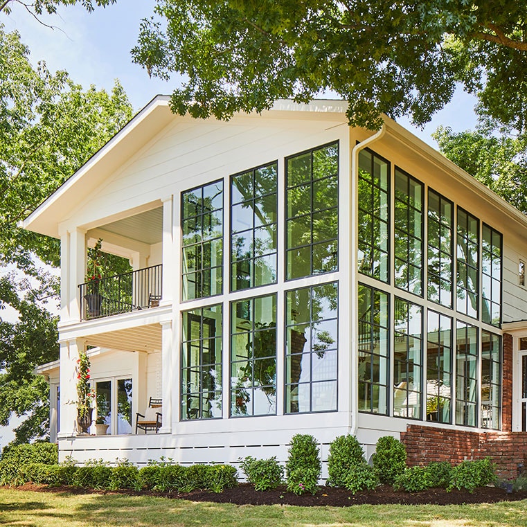 A modern house exterior features expansive floor-to-ceiling windows surrounded by greenery and trees.