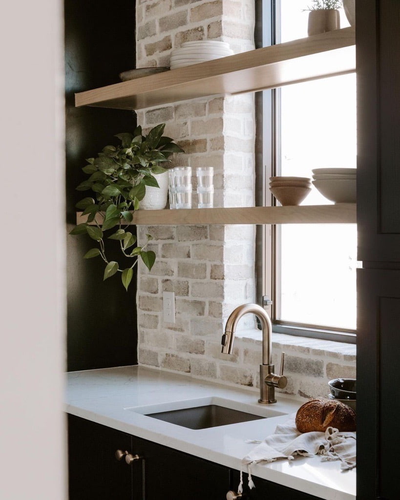 A black casement window behind the kitchen sink features shelves in front of it to display kitchen supplies.