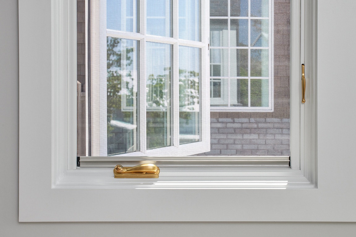 A close-up view of a casement window with a white frame and a gold handle, showcasing the outdoor scenery.