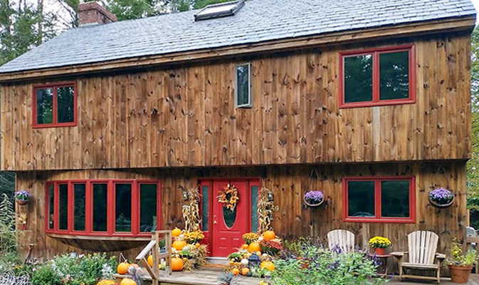 A wooden house with red windows and a red door, decorated with fall pumpkins and flowers.