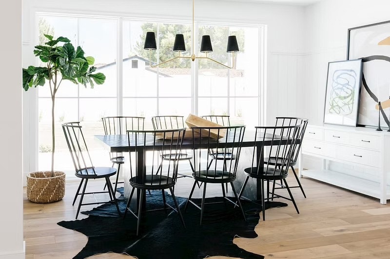 Beyond the black dining room table is a wall of white picture windows with grilles.