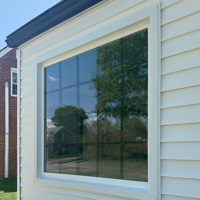 A large reflective window on a house with white siding, showing the reflection of trees and a neighboring house