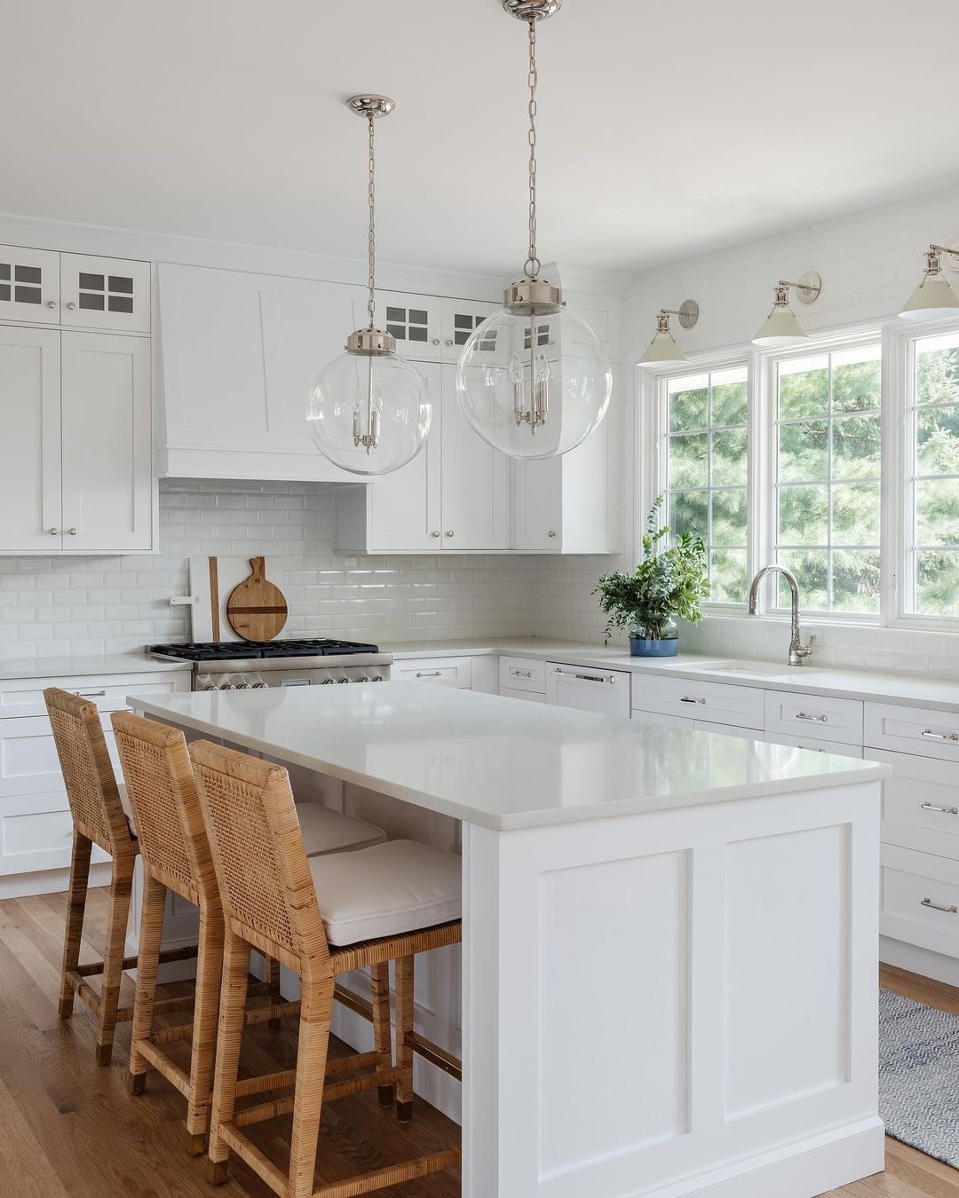 An all-white kitchen has three windows behind the sink, each with a sconce light above.