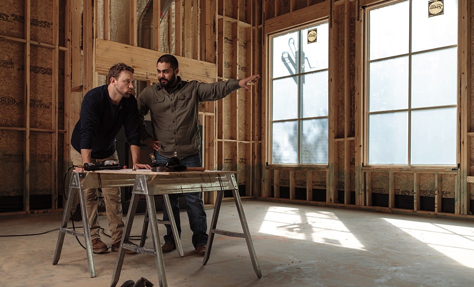 two tradesmen are looking at blueprints inside a home that is under construction.