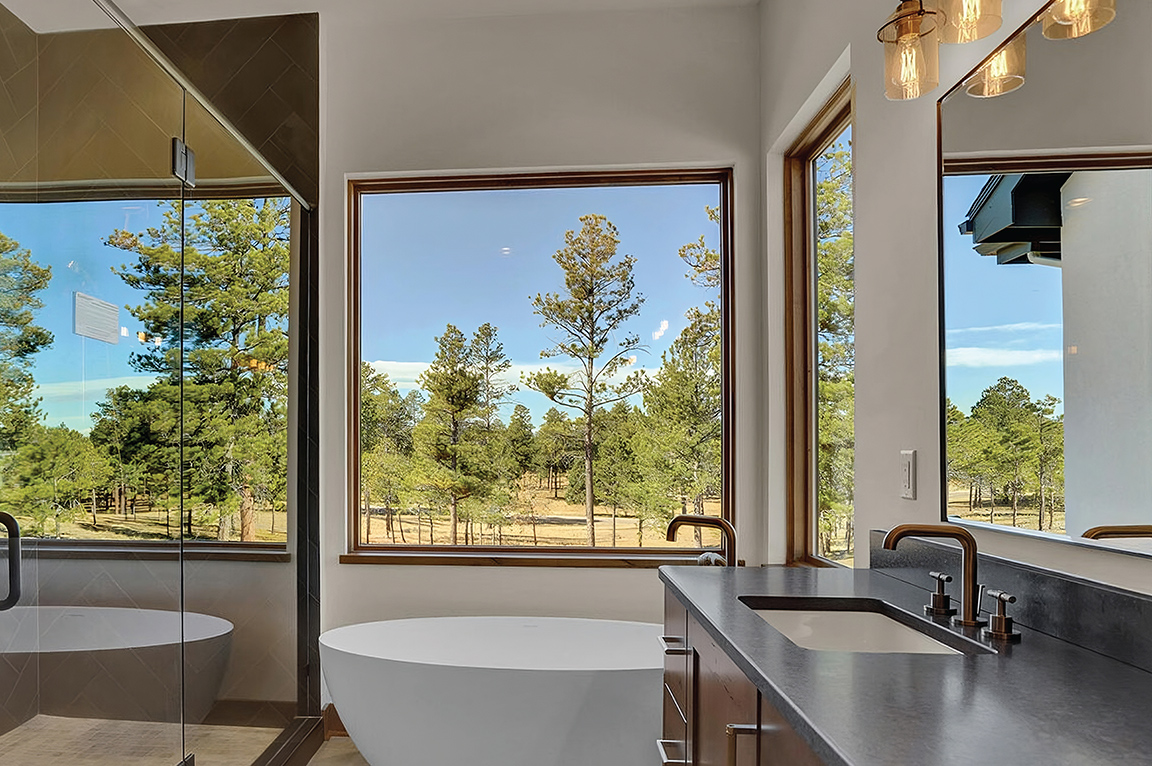 A white soaker tub sits below a large Pella wood picture window with the mountain landscape right outside.