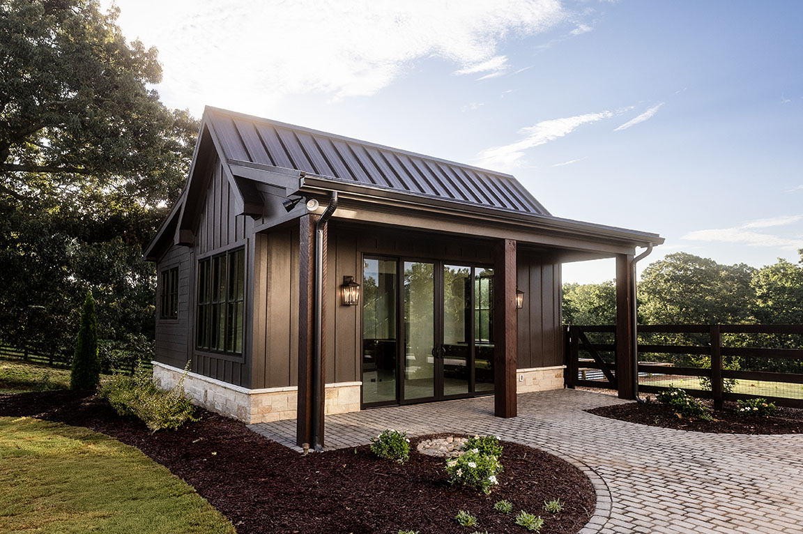 Pool house with dark siding and a metal roof, featuring large sliding glass doors.