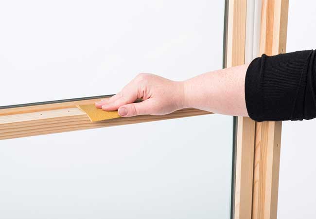 A hand holding sandpaper and sanding the wood sash of a double-hung window.