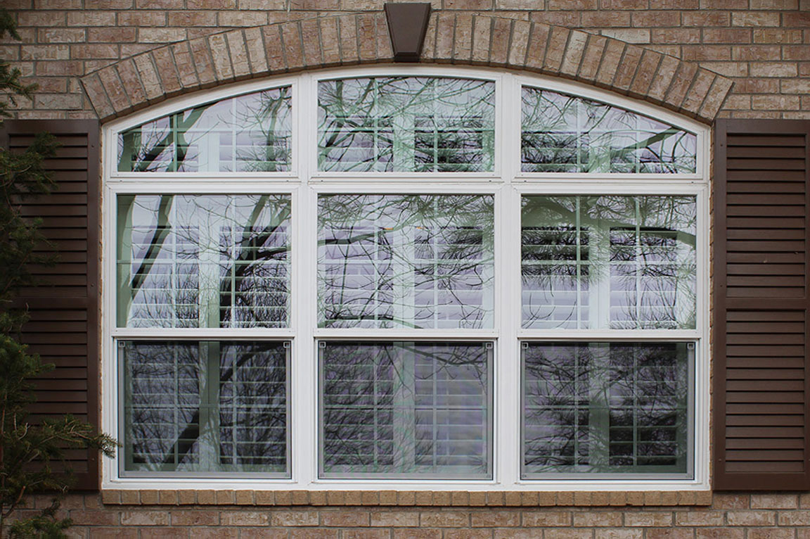 An arched vinyl window with brown shutters set against a brick wall, showcasing clear reflections.