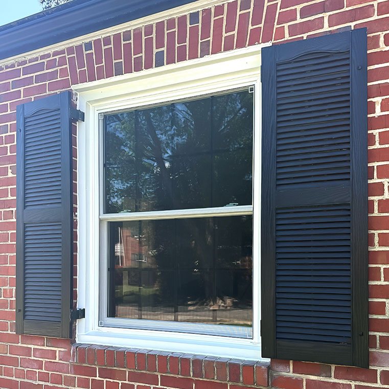 A close-up view of a window on a brick house, featuring black shutters on both sides and a white window frame