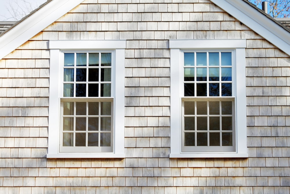 Two white double-hung windows with traditional grilles on shingle siding farmhouse