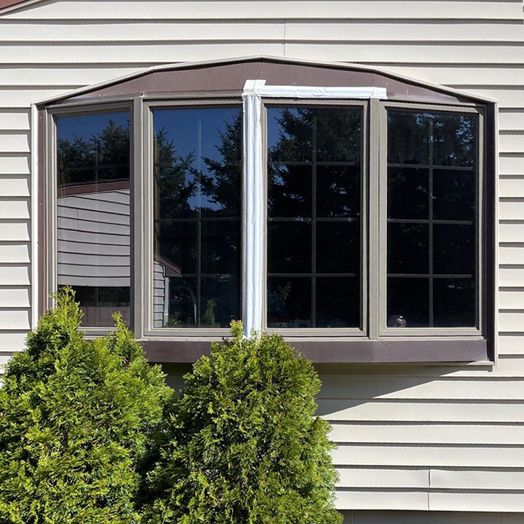 Bow window with grid pattern on tan siding, framed by greenery.