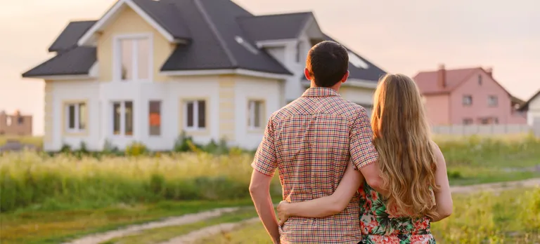 Young couple with backs to camera holding each other and looking at a house in the distance.