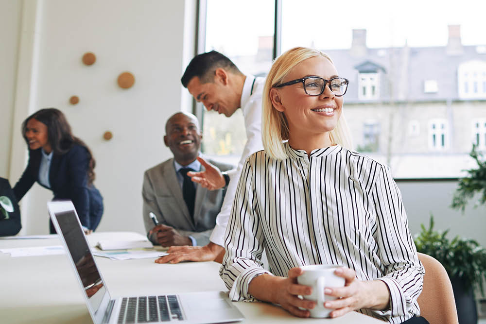 A woman sitting next to a laptop holds a coffee cup while smiling and gazing outward. Her colleagues work in the background.
