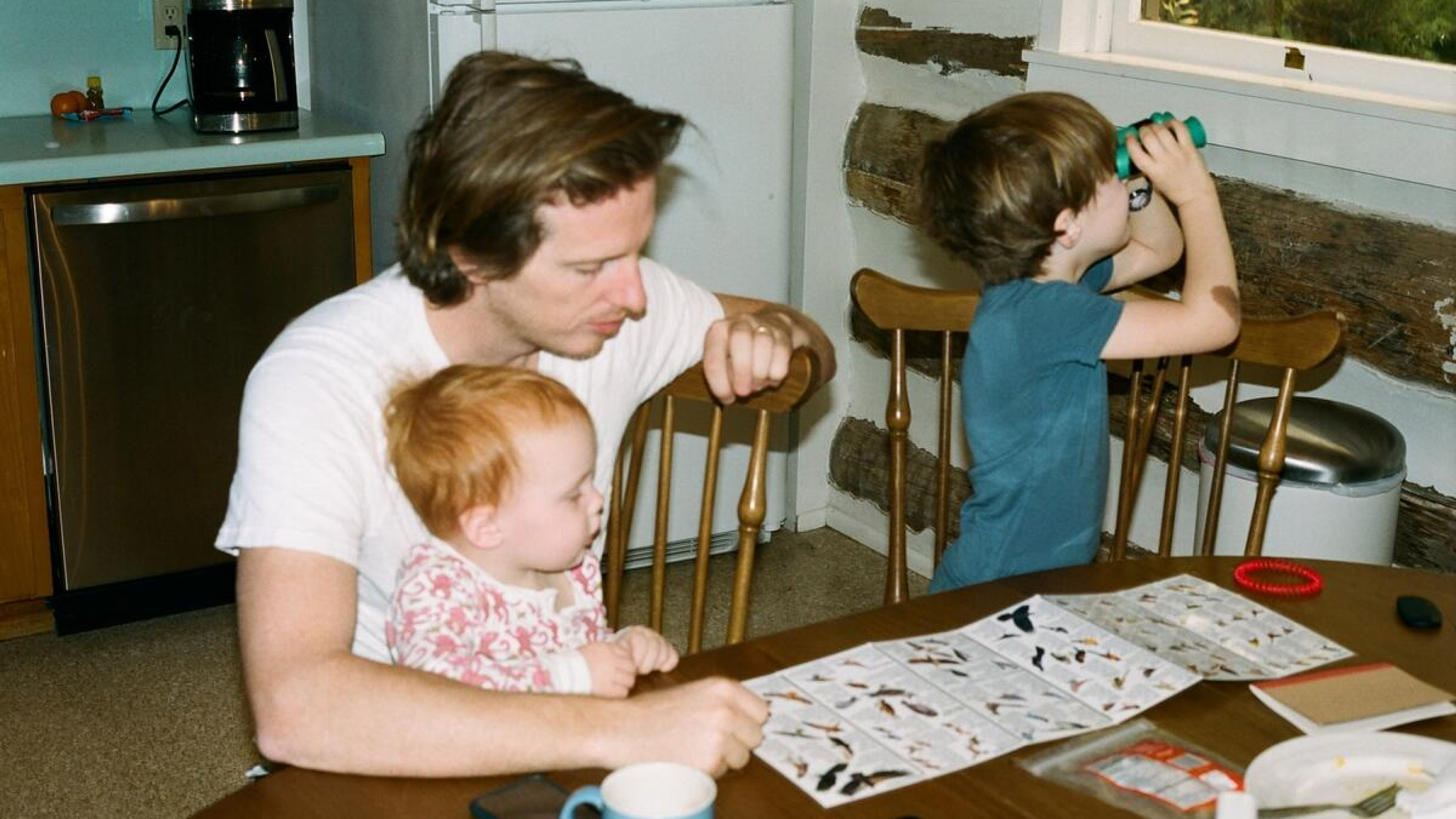 A man sits at a table with a toddler on his lap, looking at a pamphlet about birds. A young boy plays with binoculars nearby.