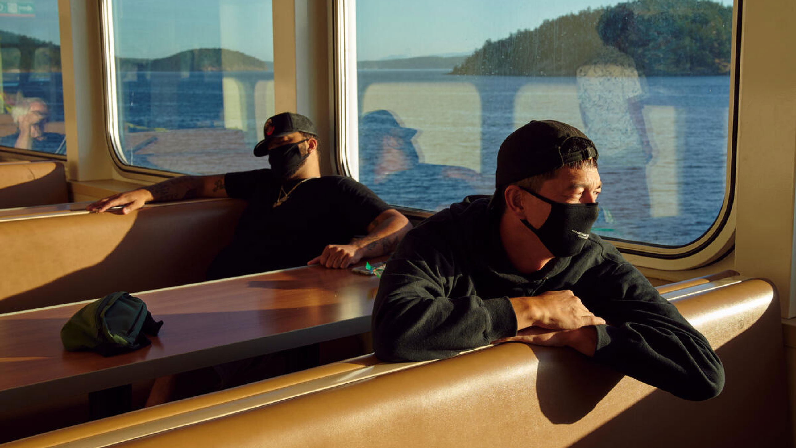 Two men sit in the sun-drenched seats of a ferry, wearing medical masks.