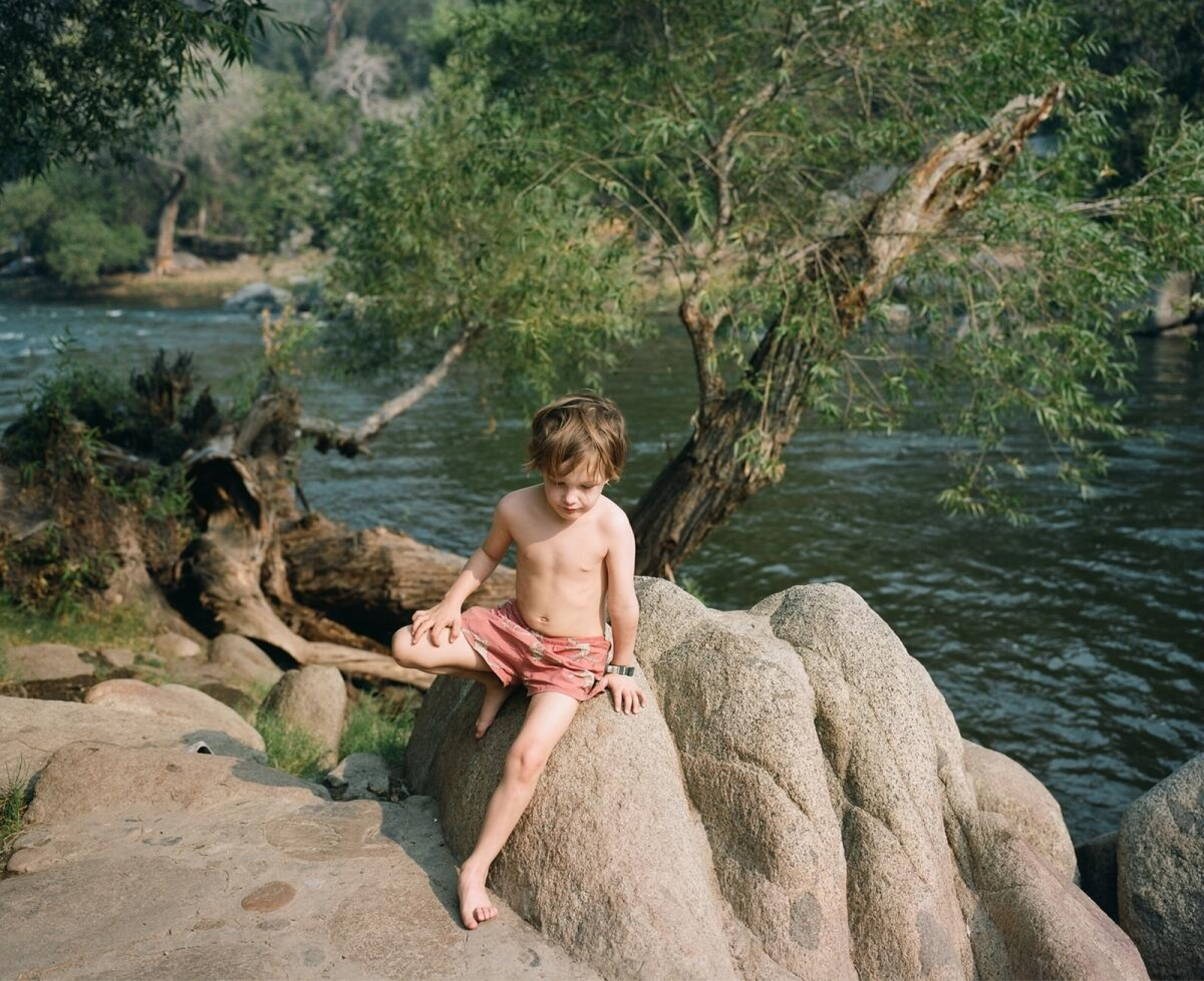 A young boy wearing swim trunks sits on a rock near a body of water.