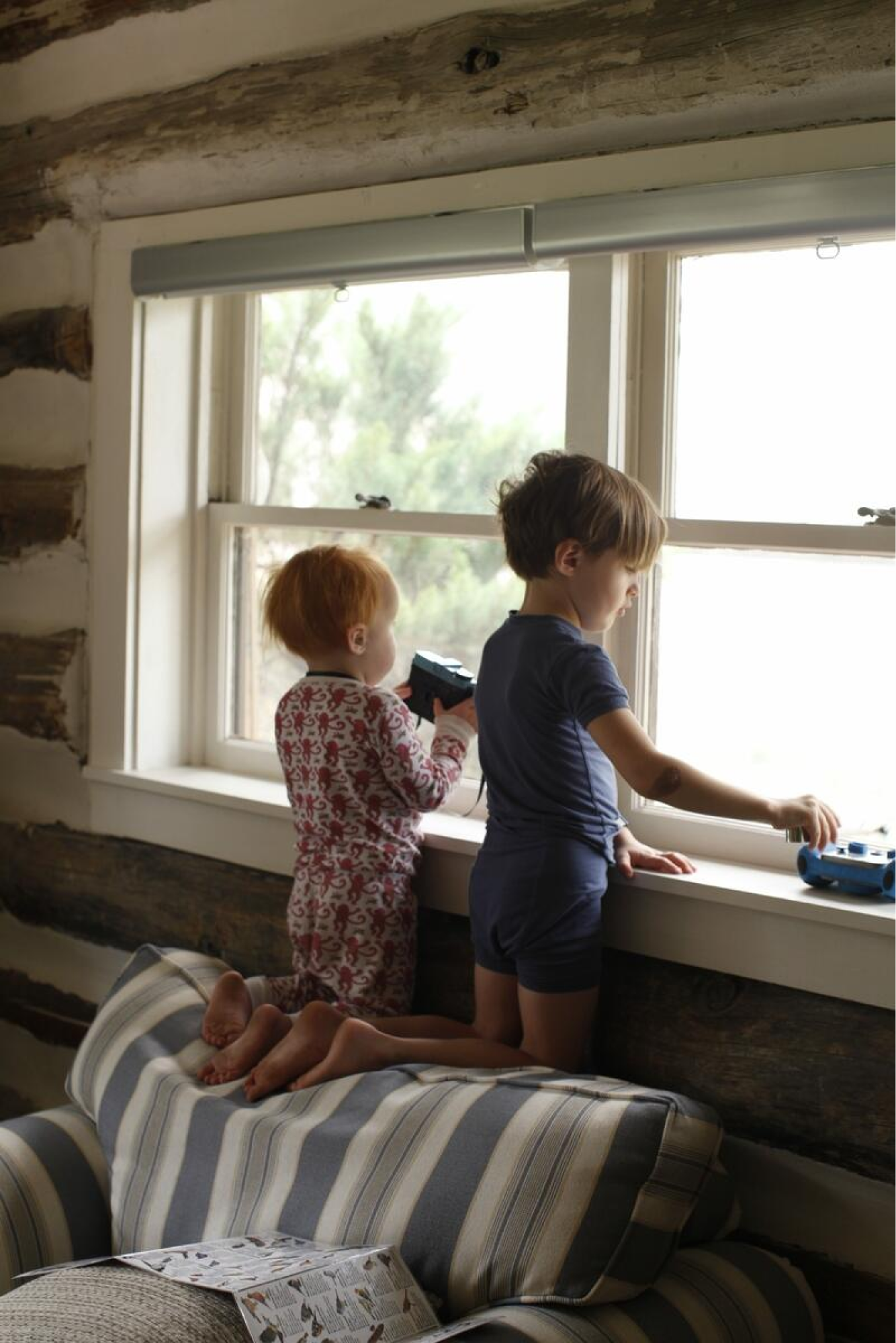 Two young children kneel on the top of a sofa to see out of a window, while one looks through binoculars.