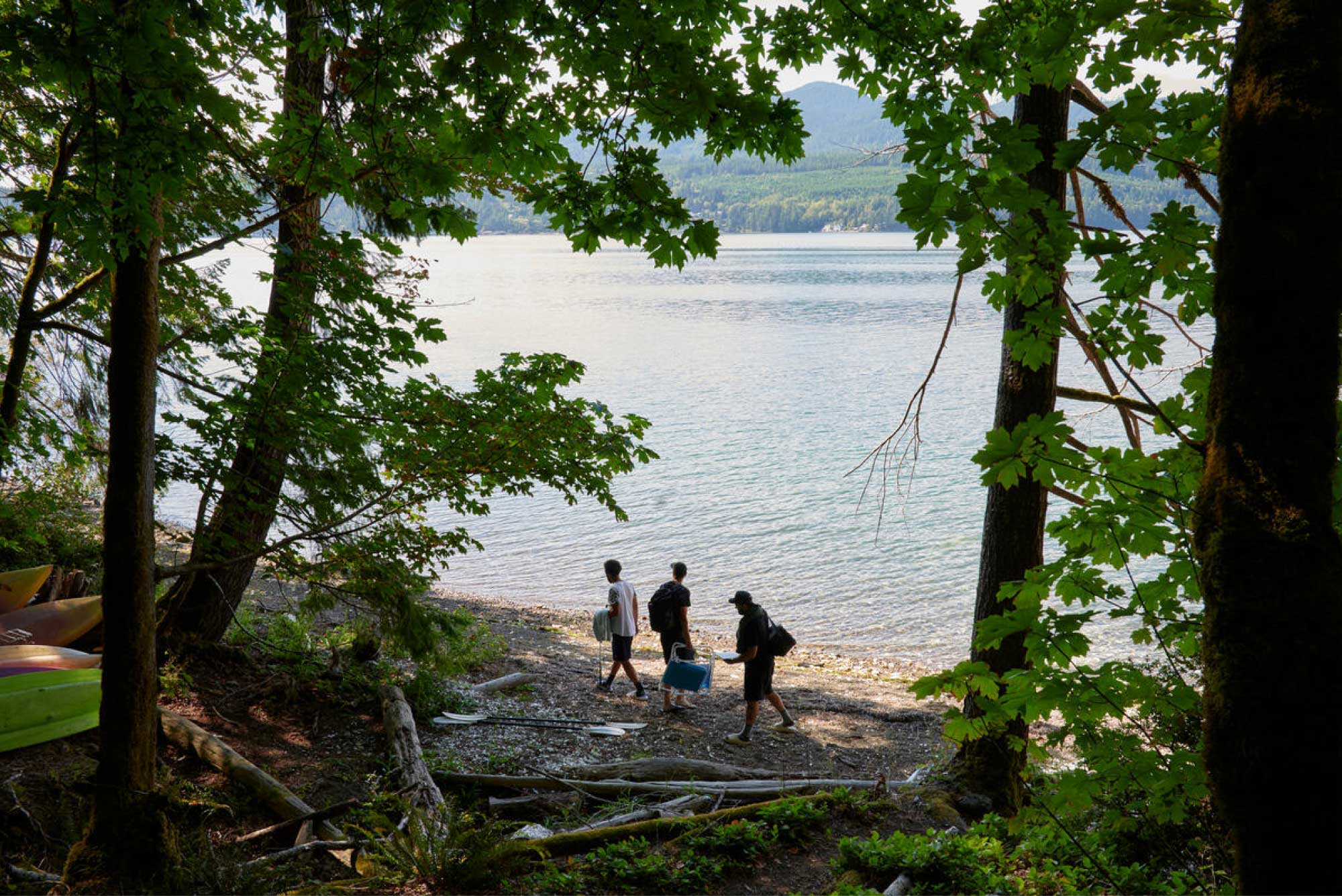 Three people stand in a forest by a body of water, holding towels and folding chairs.