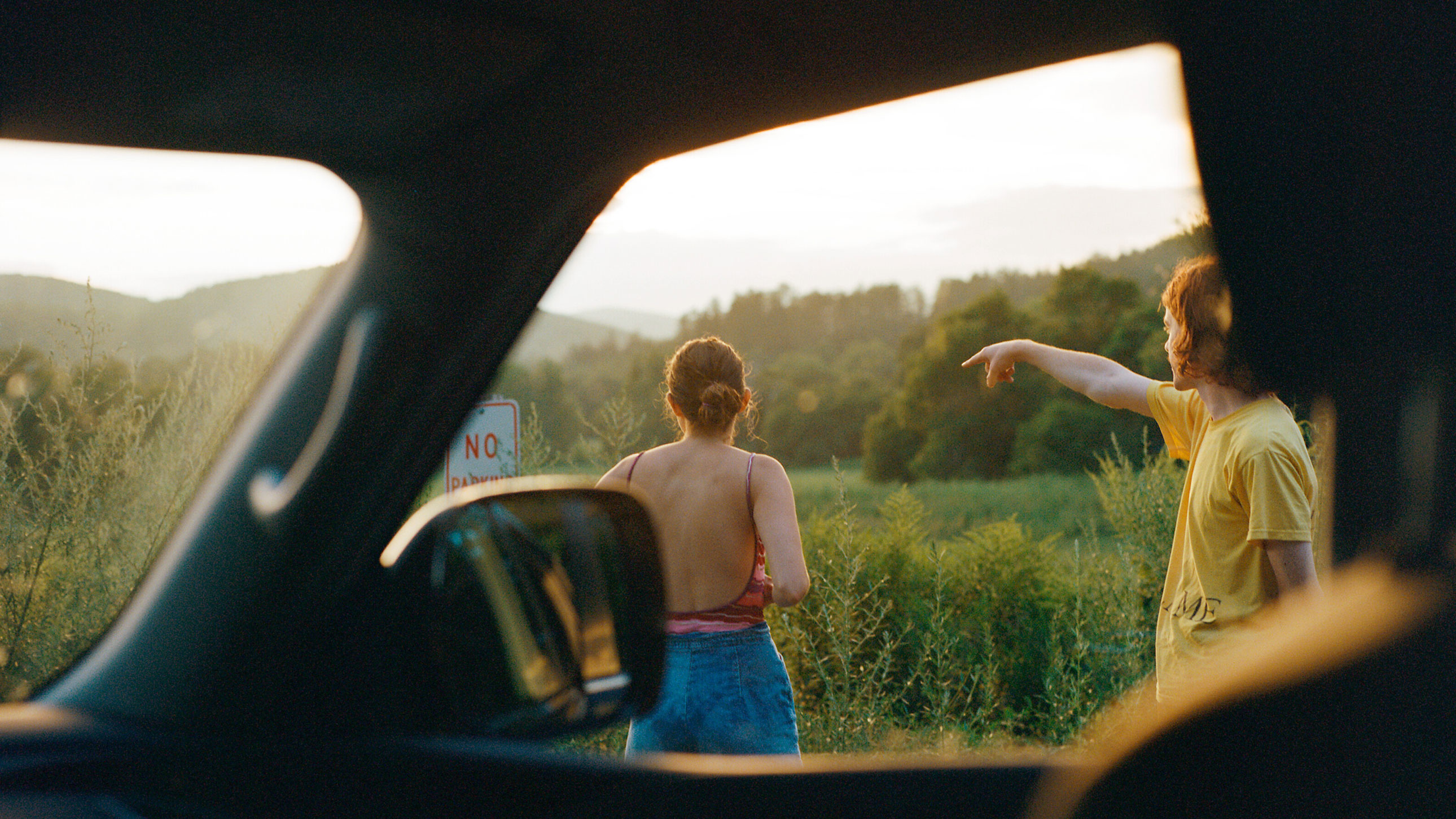 From a perspective inside of a car: two people stand outside, pointing at the rolling hills of the country side.