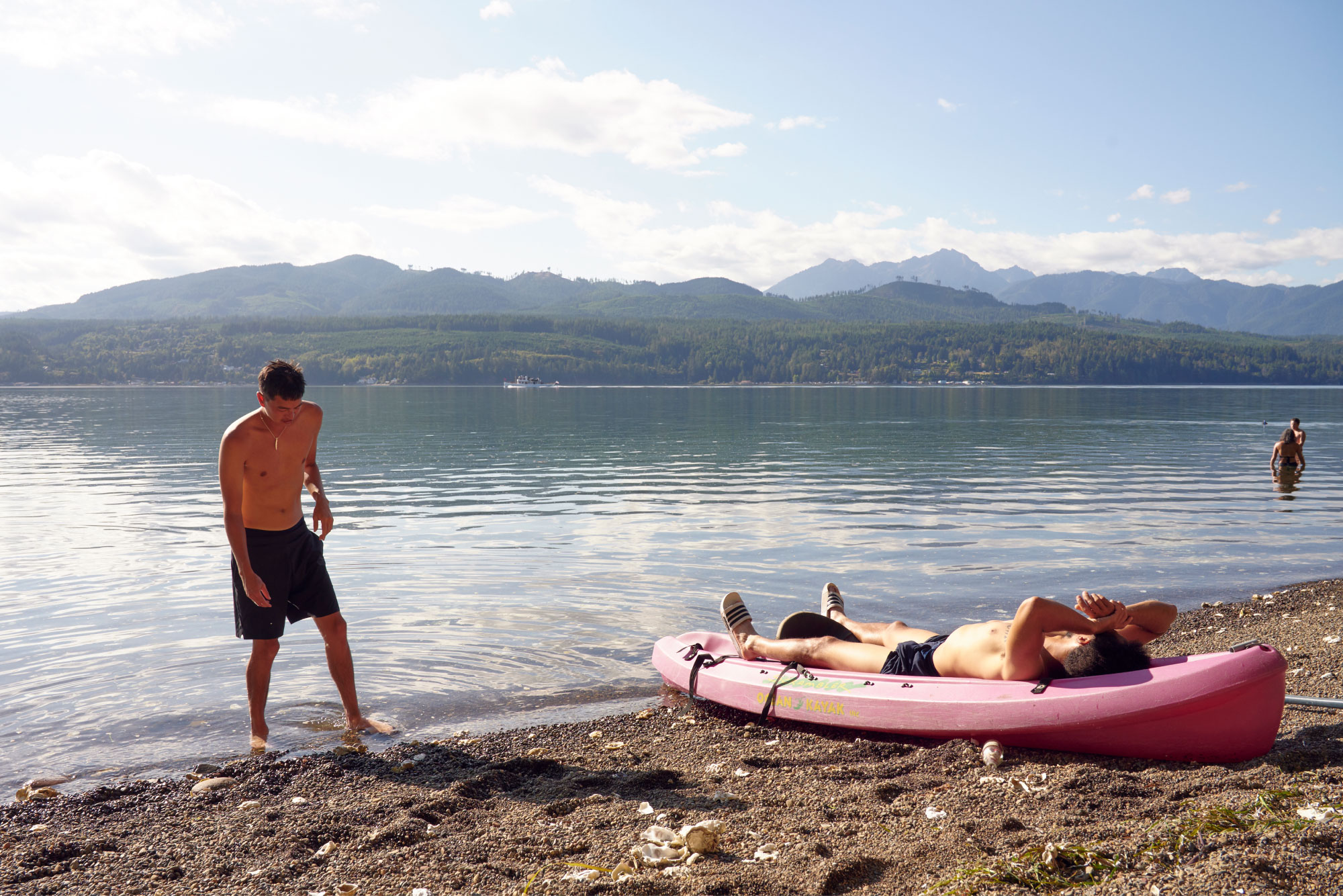 A man stands in the water by a lake, while another reclines inside of a nearby kayak.