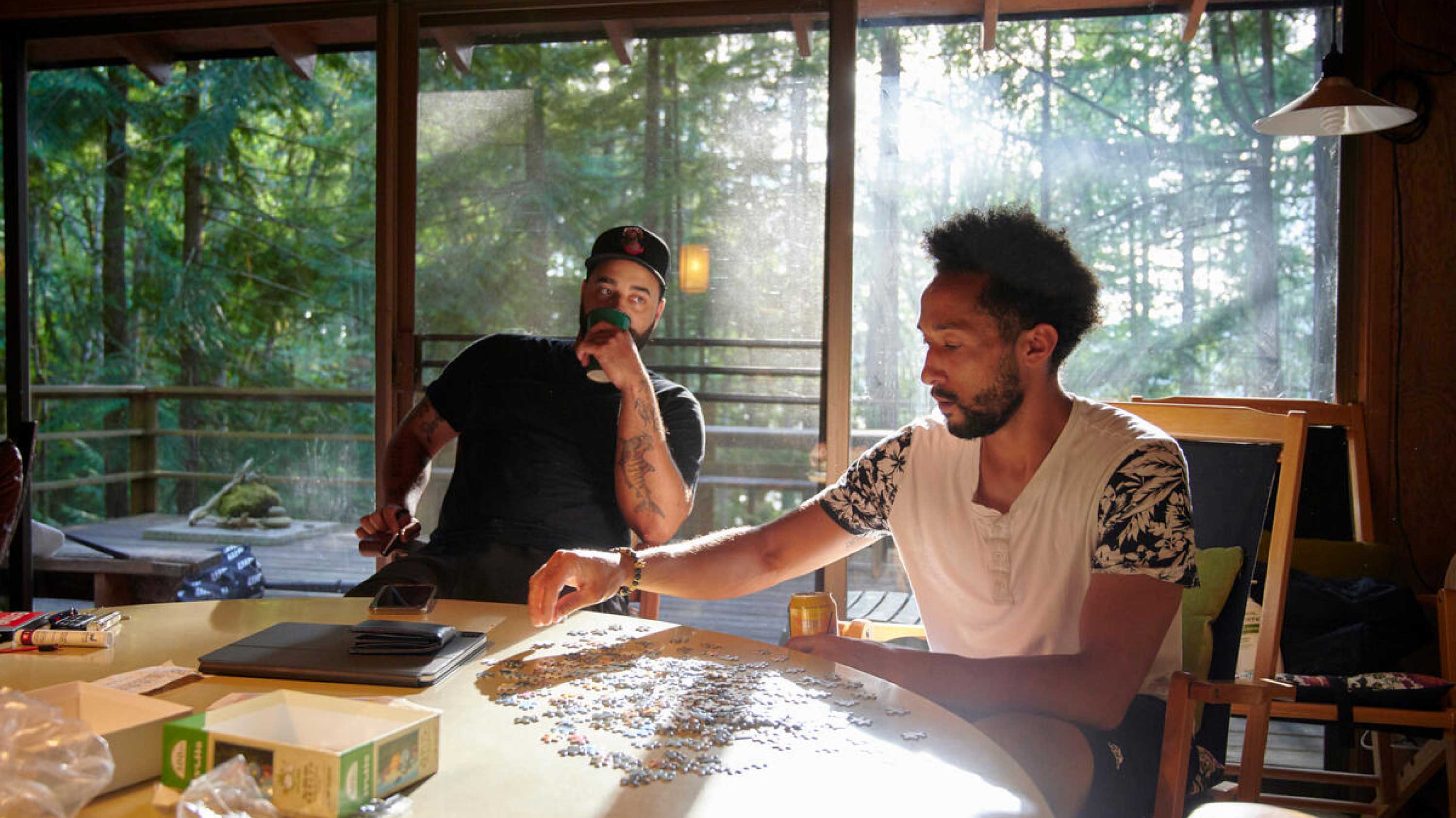 Two men sit at a table, working on a puzzle, as the sun sets through the window.