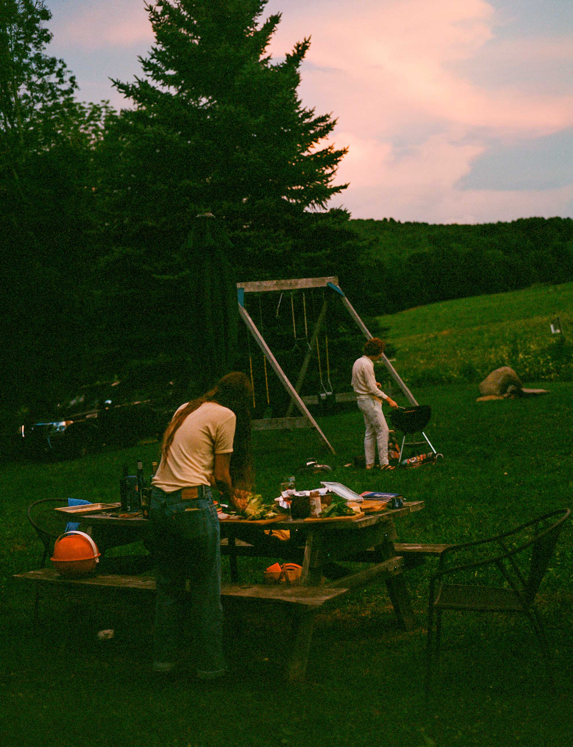 People prepare a meal outdoors at sunset, at a grill and picnic table.