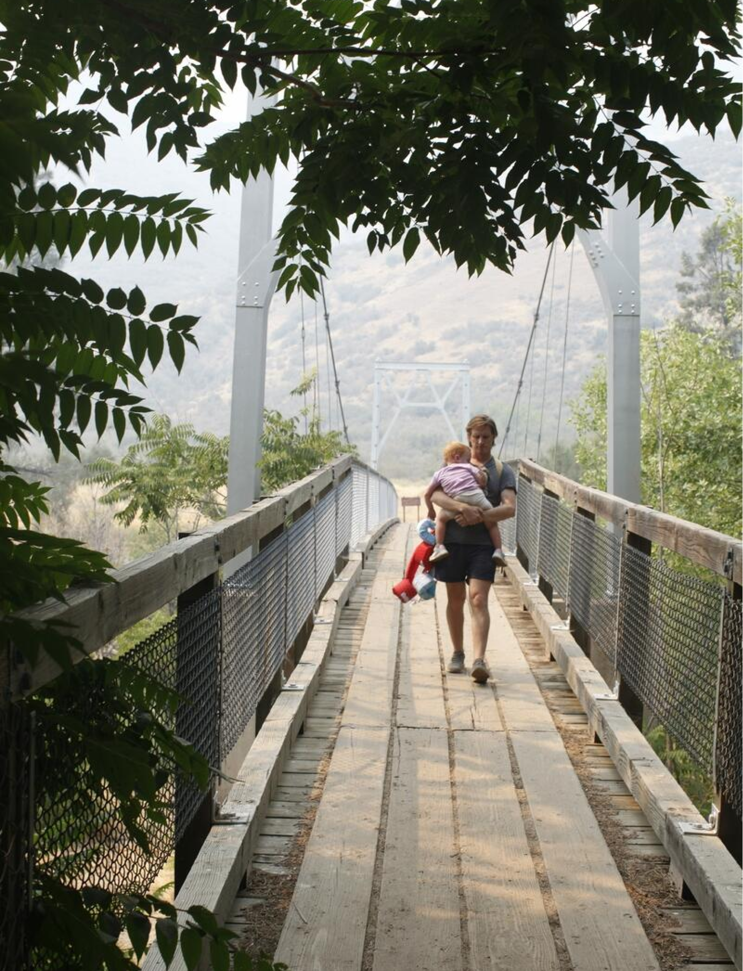 A man walks across a footbridge holding a small child in his arms.