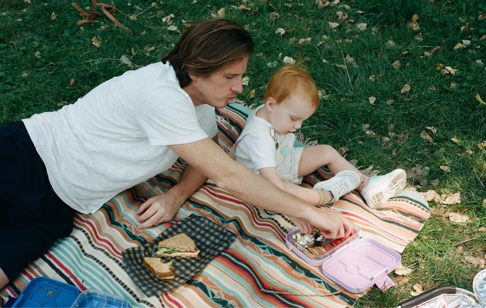 A man lies on a picnic blanket with a small child beside him and reaches for a snack.