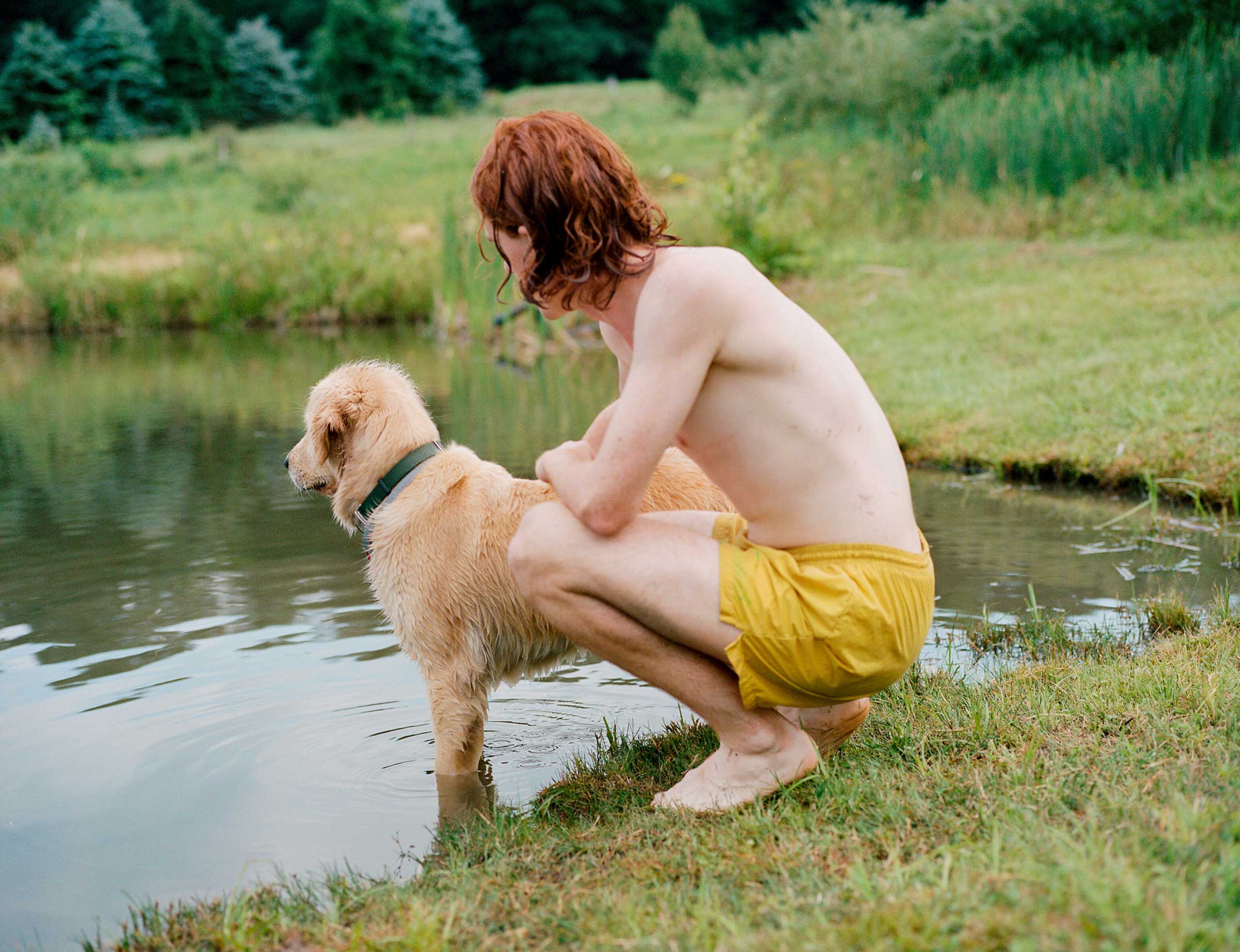 A person in swim trunks and a dog sit near a pond.