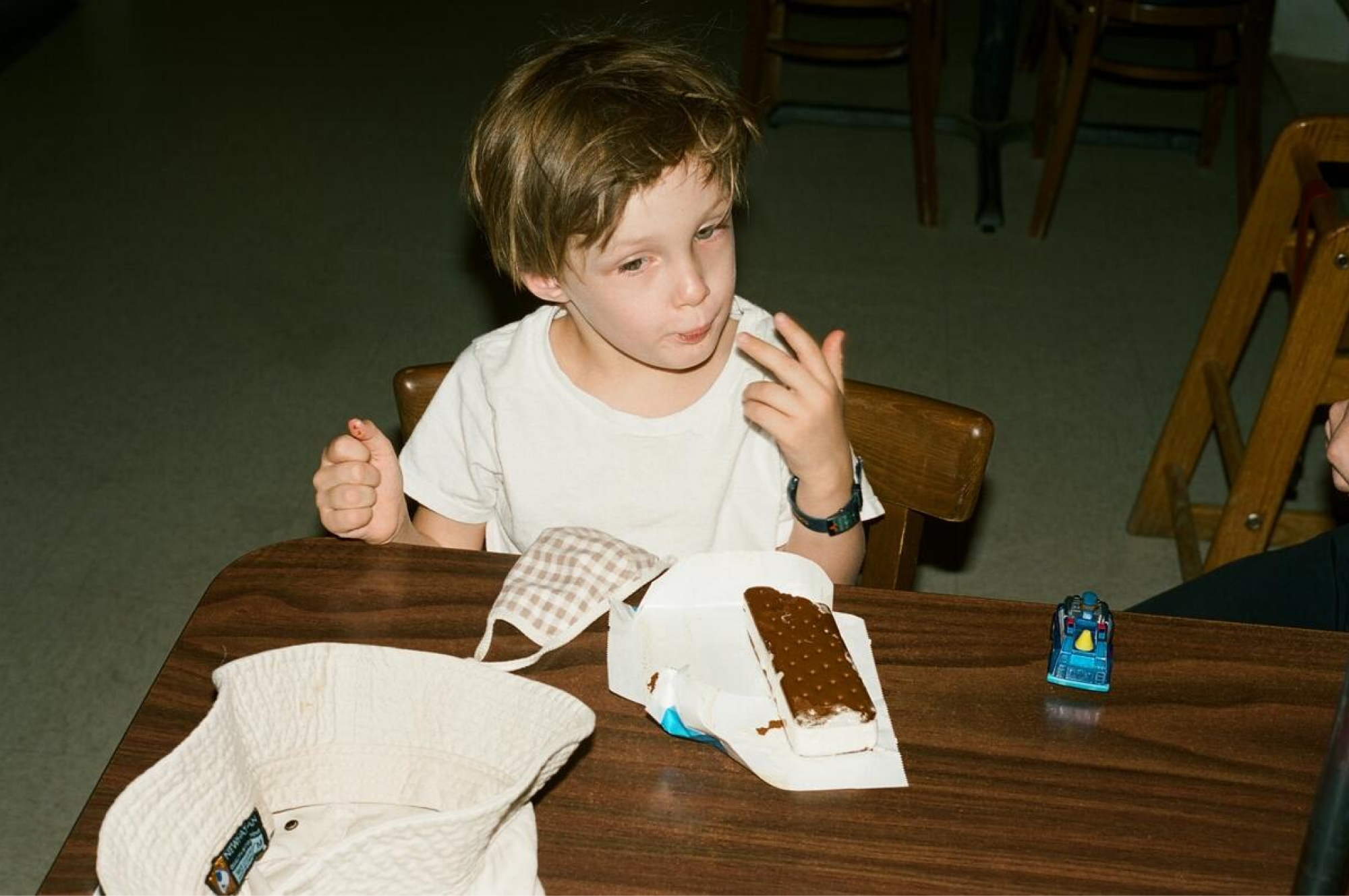 A boy sits and licks his fingers with an ice cream sandwich on the table in front of him.