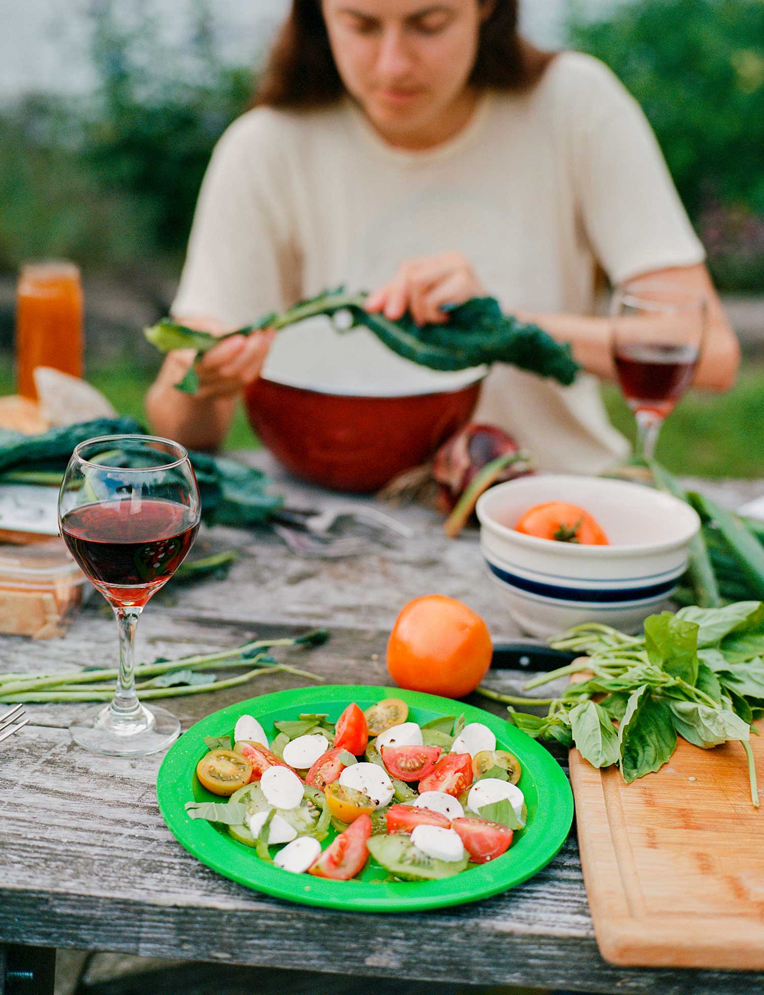 A person prepares dinner at a picnic table, near a plate with tomatoes and mozzarella.