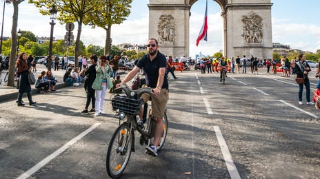 Mężczyzna jedzie na rowerze po paryskich ulicach. W tle widać łuk triumfalny.