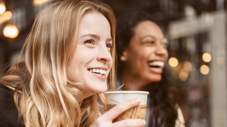 Dwie kobiety śmieją się w kawiarni. Jedna z nich pije kawę.