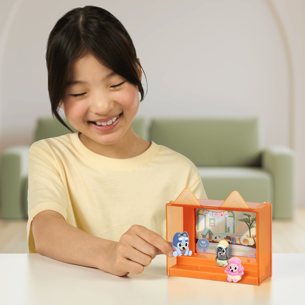Young girl sitting down, smiling, playing with 3 fuzzy Bluey characters in their display case. Display case is orange.