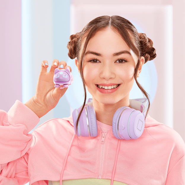 Young girl smiling at the camera holding up light purple micro pet, she is wearing headphones to match the toy.