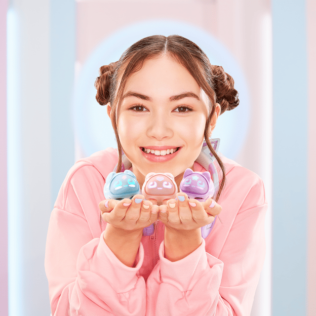 Young girl smiling at the camera holding up three different micropet toys, all different colors, under her chin.